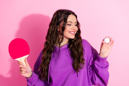 Young woman in a purple sweatshirt holding ping pong paddle and ball against pink background looking...