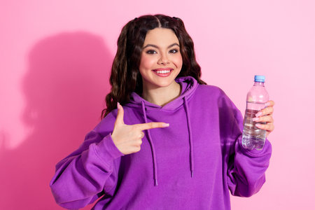 Charming young woman in a purple sweatshirt holding a water bottle and smiling, standing against a...