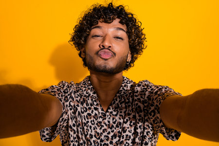 Stylish young man takes cheerful selfie striking a fun gesture against vivid yellow background showcasing trendy fashionの写真素材