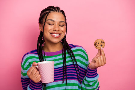 Smiling young woman wearing a colorful striped sweater holding a cookie and coffee cup on a pink backgroundの写真素材