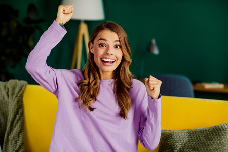 Happy young woman enjoying her time at home with joyful expressions, sitting on a cozy yellow couch in bright interiorの写真素材