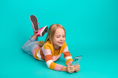 Cheerful young girl in striped sweater lying on turquoise background while using a smartphone and...