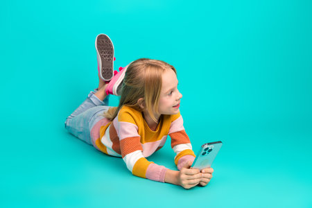 Happy young girl lying in casual attire holding smartphone on vibrant turquoise background, relaxed and cheerful poseの写真素材