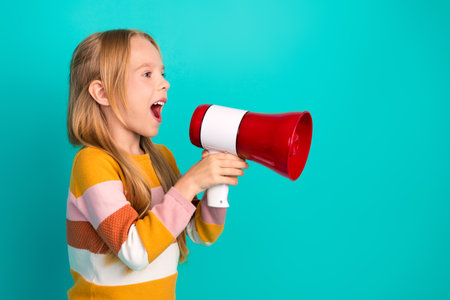 Young girl shouting through a red megaphone in front of a turquoise background, wearing a stylish...