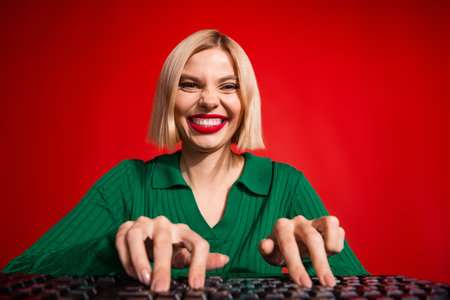 Lovely blonde woman in green sweater typing enthusiastically against vibrant red background,...