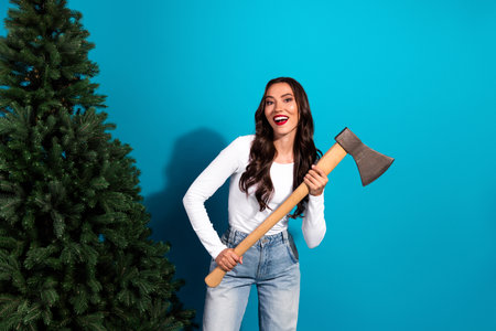 Young woman holding an axe with a Christmas tree on a blue background showcasing holiday preparation in casual attireの写真素材