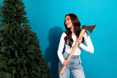 Cheerful woman holding an axe by a Christmas tree on a blue background, conveying holiday spirit and joyful preparationの写真素材