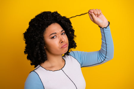 Young woman with curly hairstyle expressing puzzlement against a vibrant yellow background, showcasing casual fashion.の写真素材