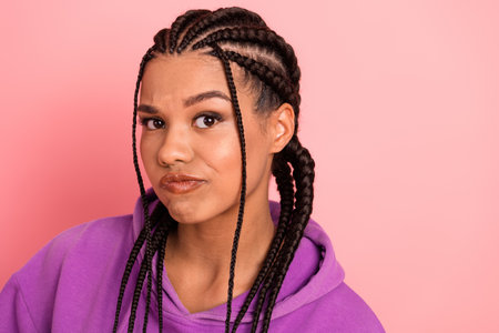Charming young woman in a braided hairstyle wearing a purple hoodie in a cheerful expression against...