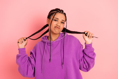 Confident young woman in stylish purple hoodie showcasing braided hairstyle with playful expression against pink backgroundの写真素材
