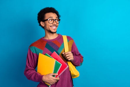 Smiling student with vibrant sweater holding notebooks on a blue background showcasing education and styleの写真素材