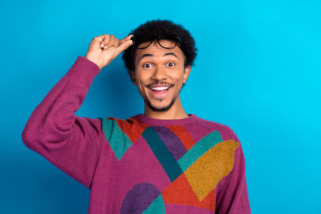 Excited young man adjusting glasses on a vibrant blue background wearing a stylish colorful sweater with a cheerful smileの写真素材