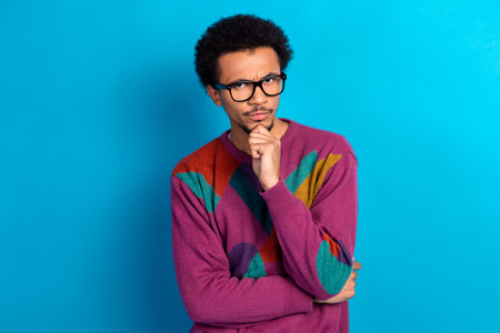 Young stylish man wearing a colorful sweater posing thoughtfully against a bright blue backgroundの写真素材