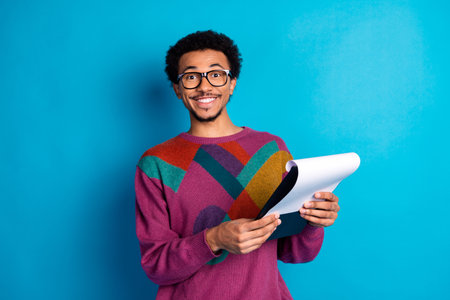 Energetic young man in modern sweater holding notebook smiling confidently against vibrant blue backgroundの写真素材