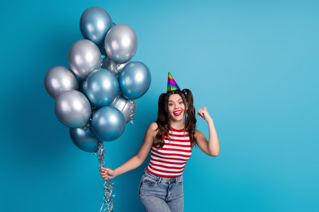 Smiling young female holding metallic balloons in party attire against a colorful background celebrating with joyの写真素材