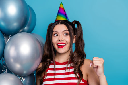 Excited young woman in striped outfit celebrating with colorful balloons and party hat against cheerful blue backgroundの写真素材