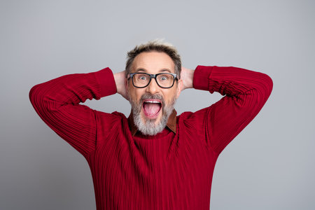 Excited middle-aged man in casual red sweater and glasses expressing astonishment on gray backgroundの写真素材