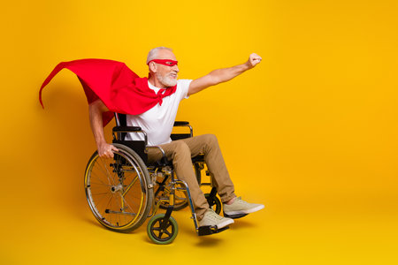 Grandpa in wheelchair wears red cape and mask and races across bright yellow studio background with joyの写真素材