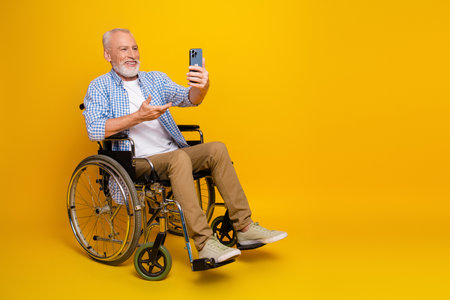 Playful grandpa in a wheelchair takes a selfie with a phone against a bright yellow background for a warm inclusive momentの写真素材
