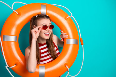 Positive woman posing playfully with lifebuoy wearing striped top and red glasses on a vibrant turquoise background.の写真素材