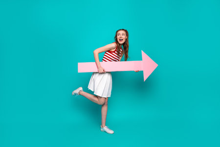 Young woman holding a pink arrow prop on teal background, emphasizing joy and creativity with trendy fashion and a cheerful poseの写真素材