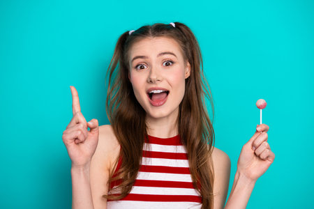 Young cheerful woman with ponytails holding a lollipop while smiling and gesturing on a vibrant backgroundの写真素材