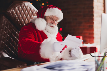 Funny Santa at his desk reading letters by the Christmas tree in a cozy home officeの写真素材