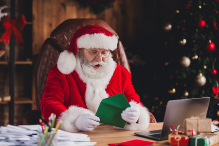 Santa at his desk opens a green card as a festive workshop scene unfolds with a Christmas tree warm lights and laptop in a cozy home office full of gifts and joyの写真素材
