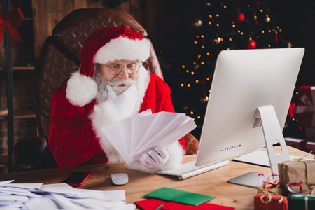 Santa Claus sits at a desk in a warm home office sorting papers beside a Christmas tree with lights and gifts creating a festive busy day vibeの写真素材
