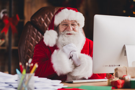 Santa at the desk laughs while planning Christmas gifts in a cozy home office by warm lights this winter eveの写真素材