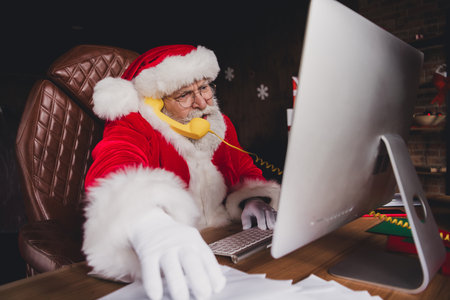 Santa at desk working on computer with yellow phone in a cozy home workshop full of holiday decorations and papers for Christmas seasonの写真素材
