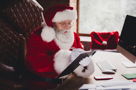 Santa at the desk creating Christmas magic with tablet and papers in a cozy home office moment full of laughter and holiday joyの写真素材