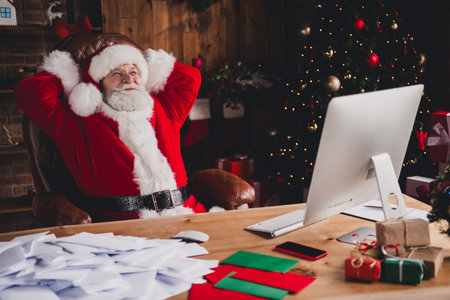 Santa at his desk relaxes at home while Christmas lights glow and a festive tree stands nearby creating a funny warm holiday scene in a cozy workshop style office setupの写真素材