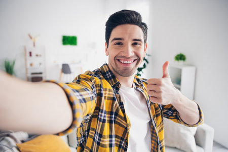 Photo of cheerful positive guy dressed plaid shirt recording video showing thumb up indoors room home houseの写真素材