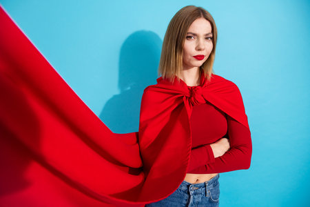 Beautiful young woman posing with confidence in a vibrant red cape and casual attire against a bright blue backgroundの写真素材