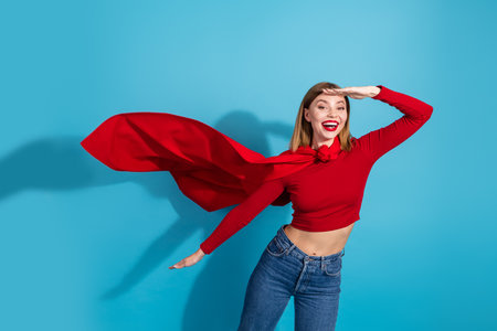 Charming young woman in casual outfit with red cape posing against blue background conveying playful and happy energyの写真素材