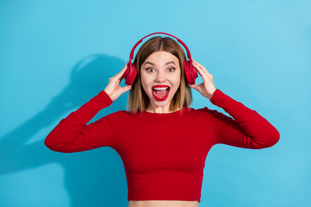 Young cheerful woman listening to music with red headphones against a bright blue background,...