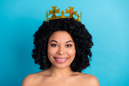 Joyful young woman with curly hair wearing a golden crown, smiling cheerfully against a bright blue backgroundの写真素材