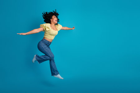 Joyful young woman jumping with arms outstretched against a vibrant blue studio background expressing carefree happinessの写真素材