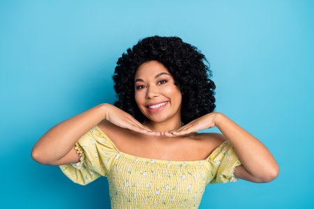 Charming young woman in yellow dress posing with her hands near face on blue background looks cheerful and happyの写真素材