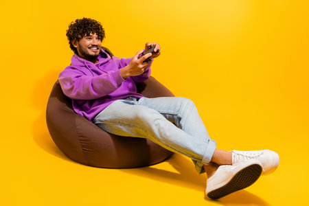 Young man enjoying gaming on a beanbag while relaxing against a bright yellow background in a vivid...