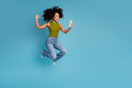 Cheerful young woman jumping in joy while looking at her smartphone against a solid blue backgroundの写真素材