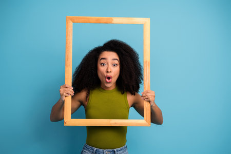 Excited young woman holding a wooden frame against a blue background, expressing surprise and charm in a casual outfitの写真素材
