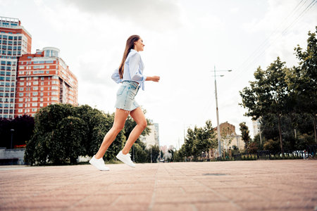 Lovely woman running outdoors in casual clothing with scenic cityscape backdrop featuring urban greeneryの写真素材