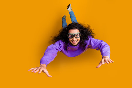 Joyful young man with curly hair and glasses posing against an orange background wearing a purple hoodie and smiling widelyの写真素材