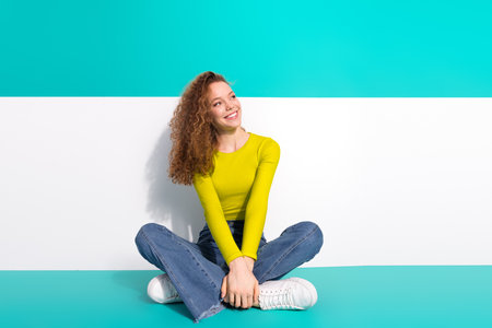 Smiling young woman in casual outfit sitting against vibrant turquoise and white background conveying cheerful and trendy atmosphereの写真素材