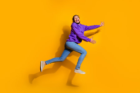 Energetic young man jumping joyfully in purple hoodie and jeans against bright yellow background...