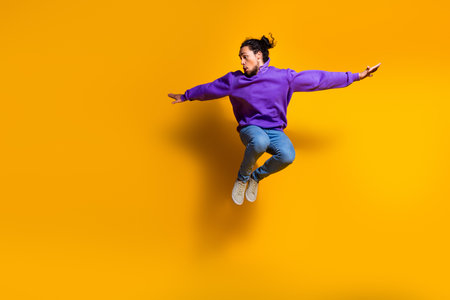 Dynamic young man wearing vibrant purple hoodie jumping in midair on a yellow background, expressing...