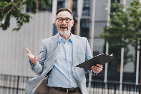 Friendly businessman in a modern city smiles and gestures while presenting a clipboard outside a sleek office building in a stylish light blue suit for stock photography listingの写真素材