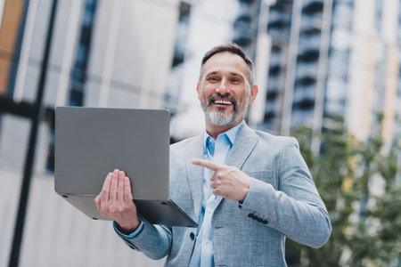 Businessman with laptop in city office environment smiles and points at screen to highlight success and modern professional lifestyle in urban megalopolis settingの写真素材